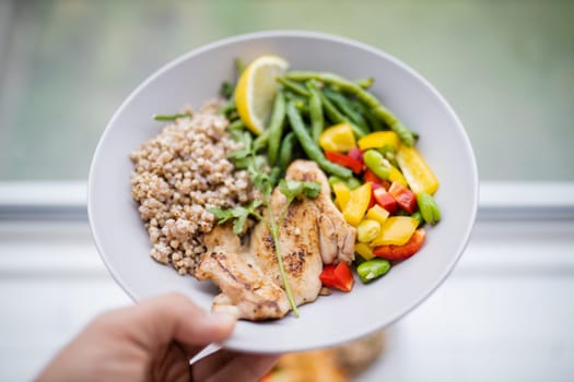 Hand holding chicken and buckwheat dish with green beans, broad beans, tomato, and pepper slices. Nutritious dish with vegetables and meat on white plate. Healthy balanced diet