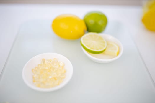 Bowl of transparent vitamins next to bowl of lemon and lime slices on white table. Medicine and citric fruit above transparent cutting board. Vitamin C nutrition