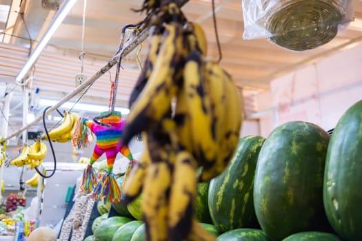 Bunch of hanging black-spotted bananas with blurry watermelons and a pinata as background. Fresh fruit with black spots for sale in classic Mexican market. Healthy food