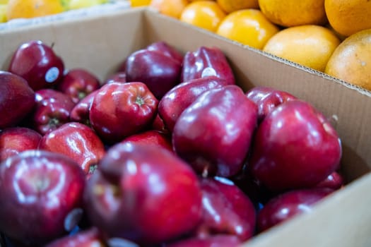 Close-up of fresh red apples in cardboard box next to pile of oranges. Juicy-looking fruit for sale on stand inside classic Mexican market. Natural healthy food