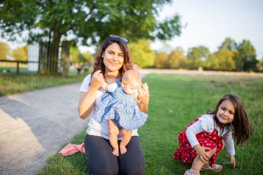 Happy woman, baby, and little girl sitting on grass with trees as background. Smiling mother playing with adorable daughters in park. Young family having fun outdoors