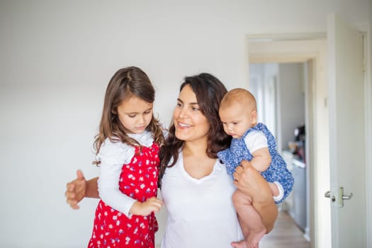Happy woman inside white bedroom lovingly hugging her adorable young daughters. Smiling mother hugging cute little girl and baby. Young family at home