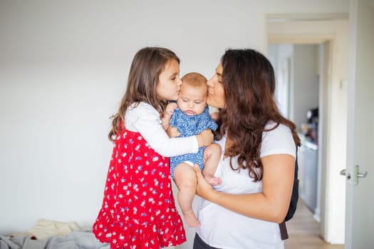 Happy woman and little girl lovingly holding and kissing adorable baby inside white room. Smiling mother and cute young daughter hugging baby. Young family at home