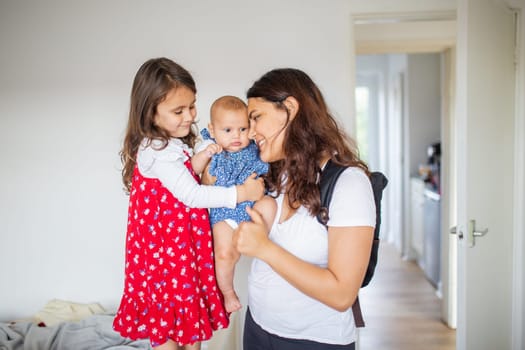 Happy woman and little girl lovingly holding adorable baby inside white room. Smiling mother and cute young daughter hugging baby. Young family at home