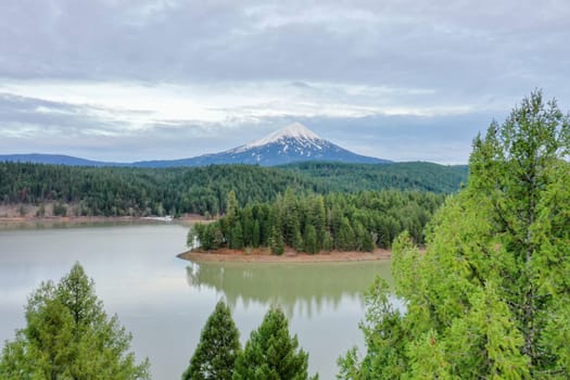 Peaceful lake in the forest with snowy mountain as background and under cloudy sky. Beautiful aerial view of lake surrounded by pine trees under cloudy skyline. Wooded and rocky landscapes
