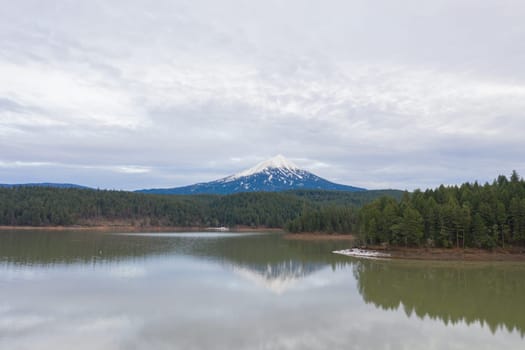 Peaceful lake in the forest with snowy mountain as background and under cloudy sky. Beautiful aerial view of lake surrounded by pine trees under cloudy skyline. Wooded and rocky landscapes