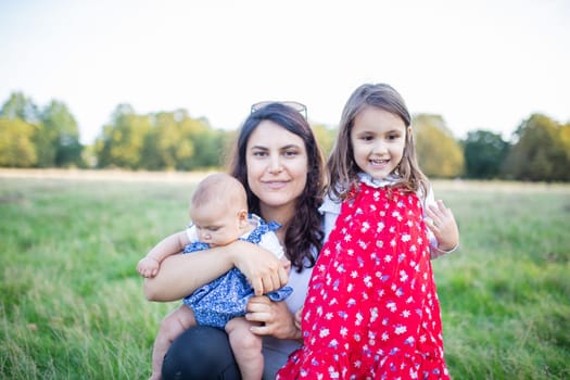 Happy woman sitting on grass with cute baby and young girl with trees as background. Mother hugging adorable daughters in park. Young family having fun outdoors