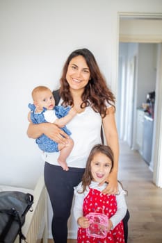 Happy mother holding adorable baby and standing next to cute young daughter. Portrait of smiling woman holding baby and young child. Lovely young family at home