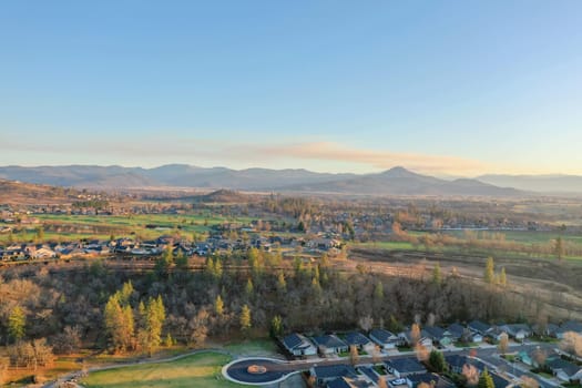 Peaceful town surrounded by trees with mountains as background during sunset. Aerial view of roads and houses surrounded by autumn-colored nature. Rural town landscape