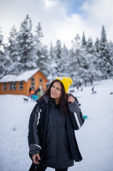 Happy woman standing with a beautiful snowy forest and small cabin as background. Portrait of smiling woman surrounded by snowy pine trees. Adventurous winter holiday