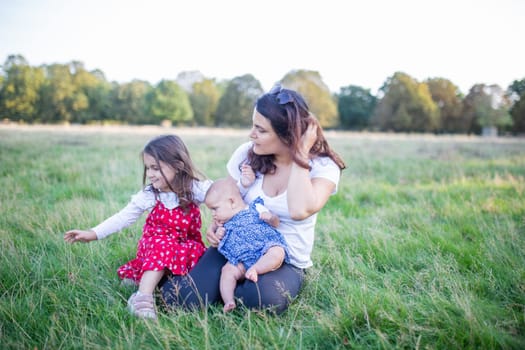 Happy woman, baby, and little girl sitting on grass with trees as background. Mother playing with adorable young daughters in park. Young family having fun outdoors