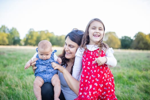 Happy woman, baby, and little girl sitting on grass with trees as background. Smiling mother holding adorable daughters in park. Young family having fun outdoors