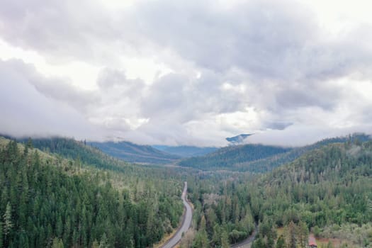 Beautiful and peaceful forest under cloudy sky and with mountains as background. Aerial view of road in the middle of the woods. Wooded and rocky landscapes