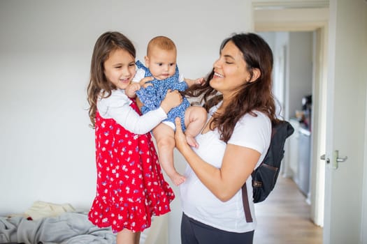 Happy woman and little girl lovingly holding adorable baby inside white room. Smiling mother and cute young daughter hugging baby. Young family at home