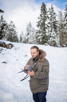 Handsome man holding drone with snowy pines as background. Portrait of concentrated man interacting with drone in frozen woods. Adventurous winter holiday