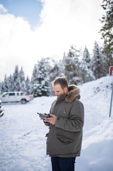 Handsome man holding drone remote control with a beautiful snowy forest as background. Portrait of concentrated man controlling drone in frozen woods. Adventurous winter holiday