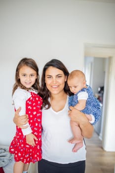 Happy woman inside white bedroom lovingly hugging her adorable young daughters. Smiling mother tightly holding cute little girl and baby. Young family at home