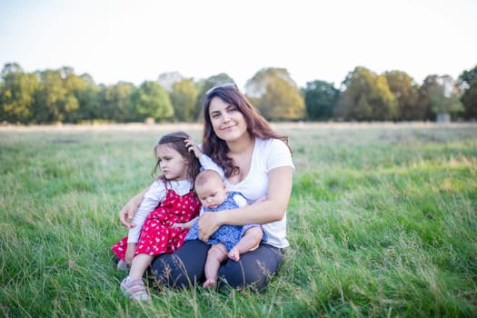 Happy woman, baby, and little girl sitting on grass with trees as background. Smiling mother hugging adorable daughters in park. Young family having fun outdoors