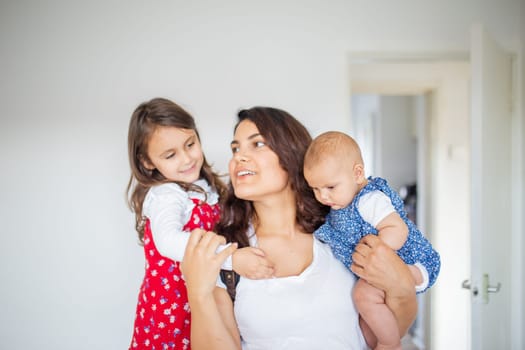 Happy woman inside white bedroom lovingly hugging her adorable young daughters. Smiling mother hugging cute little girl and baby. Young family at home
