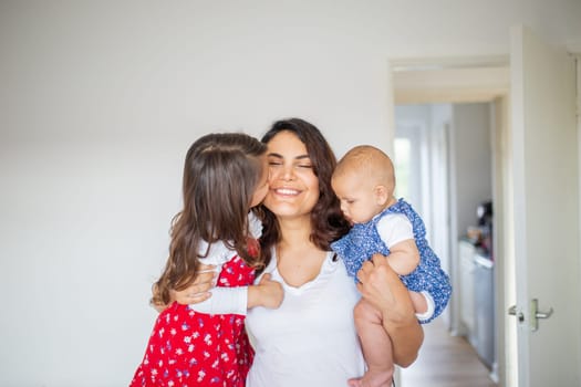 Little girl lovingly hugging and kissing her happy mother while she holds cute baby.Smiling mother hugging cute little girl and baby. Young family at home