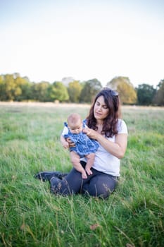 Happy woman sitting on grass and holding her cute baby with trees as background. Mother and daughter relaxing in park under clear sky. Young family having fun outdoors