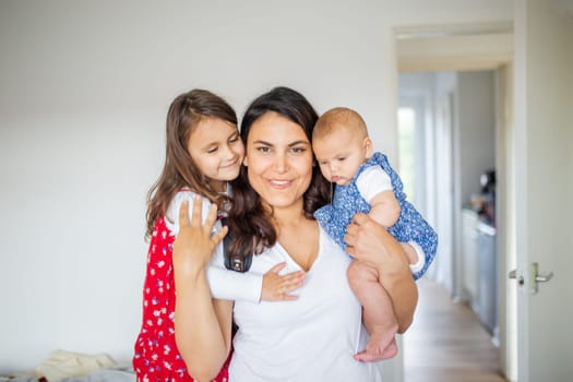 Happy woman inside white bedroom lovingly hugging her adorable young daughters. Smiling mother hugging cute little girl and baby. Young family at home