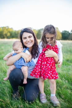 Happy woman sitting on grass with cute baby and young girl with trees as background. Smiling mother holding adorable daughters in park. Young family having fun outdoors