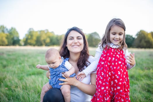 Happy woman sitting on grass with cute baby and young girl with trees as background. Smiling mother holding adorable daughters in park. Young family having fun outdoors