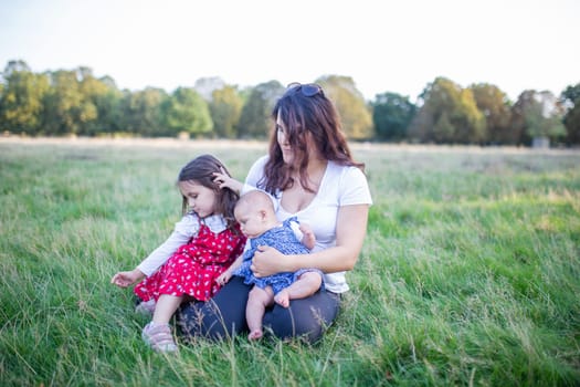 Happy woman, baby, and little girl sitting on grass with trees as background. Mother playing with adorable young daughters in park. Young family having fun outdoors