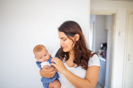 Woman lovingly holding her adorable baby inside white bedroom. Portrait of young mother hugging cute baby daughter. Young family at home