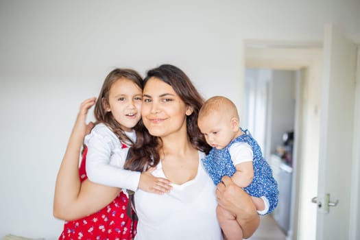 Happy woman inside white bedroom lovingly hugging her adorable young daughters. Smiling mother hugging cute little girl and baby. Young family at home