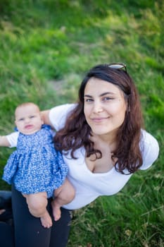 Beautiful smiling woman sitting on grass with her cute baby. Portrait of joyful mother and daughter relaxing in park. Young family having fun outdoors