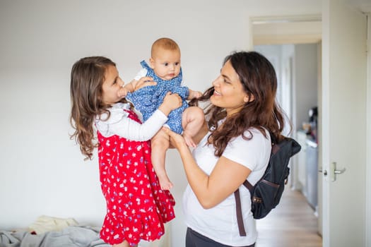 Happy woman and little girl lovingly holding adorable baby inside white room. Smiling mother and cute young daughter hugging baby. Young family at home