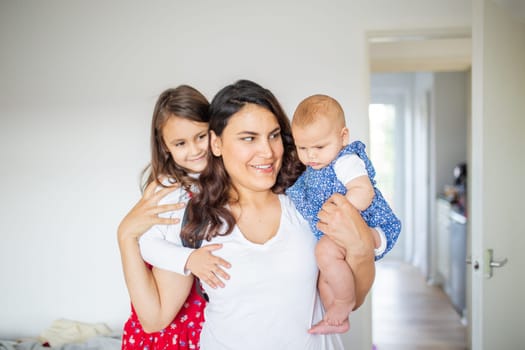 Happy woman inside white bedroom lovingly hugging her adorable young daughters. Smiling mother hugging cute little girl and baby. Young family at home