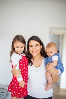 Happy woman inside white bedroom lovingly hugging her adorable young daughters. Smiling mother tightly holding cute little girl and baby. Young family at home