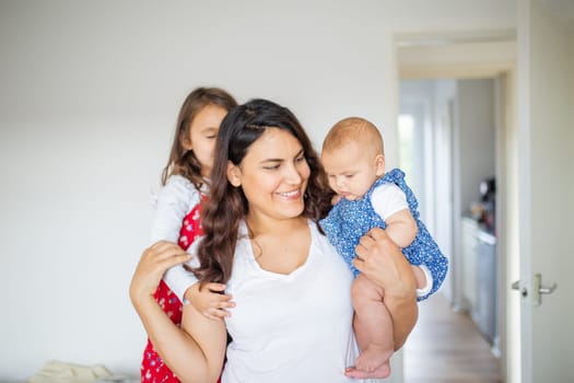 Happy woman inside white bedroom lovingly hugging her adorable young daughters. Smiling mother hugging cute little girl and baby. Young family at home