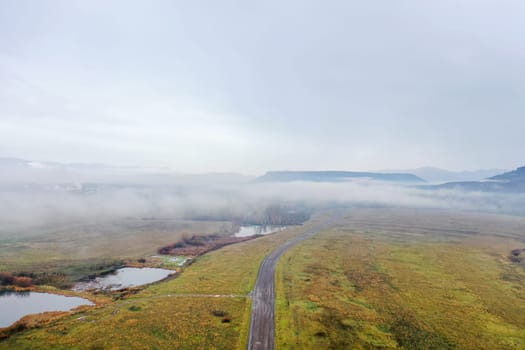 Road in the middle of open field and under cloudy sky with mountains as background. Aerial view of paved road and marsh on empty grassland. Foggy landscapes