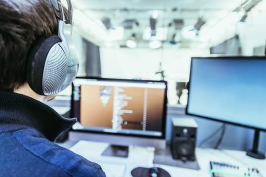 Young man with headphones is sitting in the recording studio, buttons, studio, screens and equipment in the blurry background