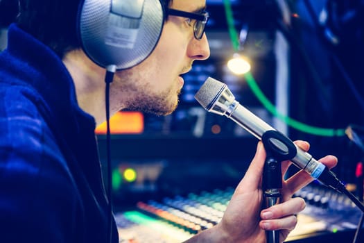 Young man is sitting in the recording studio and talks into the microphone, buttons and equipment in the blurry background