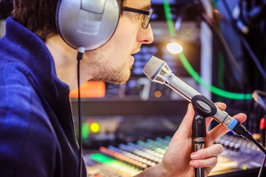 Young man is sitting in the recording studio and talks into the microphone, buttons and equipment in the blurry background