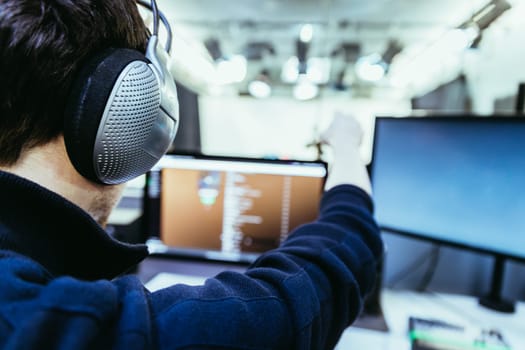 Young man with headphones is sitting in the recording studio and makes a gesture. Buttons, studio, screens and equipment in the blurry background