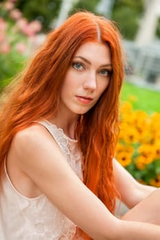 attractive young woman with long red hair sits in the garden on a wall and relaxes in summer in daylight and good weather