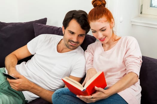 Young couple in the living room on the couch while reading and watching TV at home