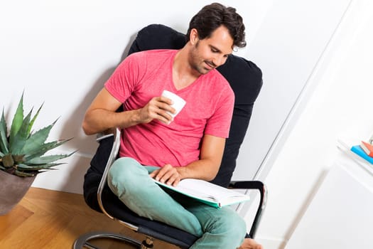 Young man in casual clothes relaxes while reading in an armchair with a drink in his hand