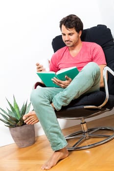 Young man in casual clothes relaxes while reading in an armchair with a drink in his hand