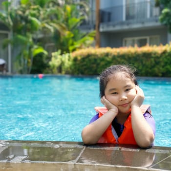 Asian child playing in the pool. Wearing orange life jacket, rests her chin on both hands by the edge of the pool.
