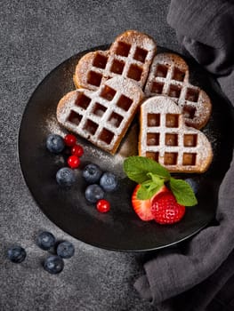 Delicious waffles heart shape with  powdered sugar and berries on ceramic black plate on grey background. Top view. Sweet meal. Dessert. Serving food