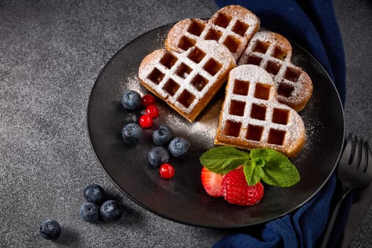 Delicious waffles heart shape with  powdered sugar and berries on ceramic black plate on grey background. Top view. Sweet meal. Dessert. Serving food