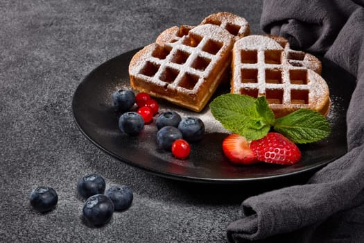 Delicious waffles heart shape with  powdered sugar and berries on ceramic black plate on grey background. Sweet meal. Dessert. Serving food