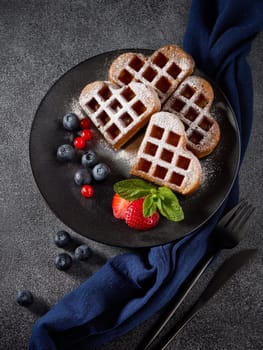 Delicious waffles heart shape with  powdered sugar and berries on ceramic black plate on grey background. Top view. Sweet meal. Dessert. Serving food